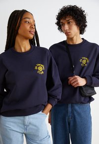 Σκούρο μπλε φούτερ με κίτρινο λογότυπο "On Vacation Club", με ριμπ μανσέτες και στρογγυλή λαιμόκοψη; συνδυασμένο με ανοιχτό μπλε και σκούρα τζιν.