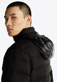 Czarna kurtka puchowa o teksturowanym połysku, pikowanym designie i ściąganym kapturze. Materiał wydaje się gładki i ocieplający dla zapewnienia ciepła.