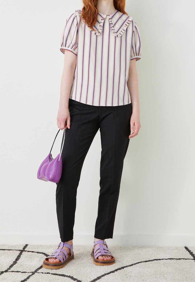 Personne portant un chemisier à manches courtes rayé avec col à volants, pantalon noir, sandales violettes et tenant un petit sac à main violet.