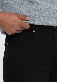 Jeans en denim noir avec une coupe slim, dotés de finitions en métal argenté, de poches avant et d'une texture subtile, portés avec un pull en tricot gris.