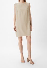 Vestido beige sin mangas con corte recto, que presenta una tela texturizada y bolsillos laterales, combinado con sandalias de tacón bloque ligero con tiras.