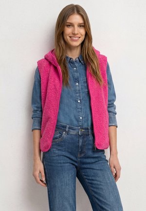 Frau lächelt, trägt blaues Jeanshemd und Jeans mit einer leuchtend pinken ärmellosen Fleecejacke, steht vor einer weißen Wand.
