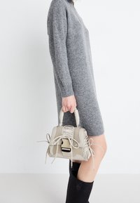 Borsa beige con dettagli intrecciati e inserti in pizzo, caratterizzata da una forma strutturata e manici corti, tenuta da una persona in un abito grigio.