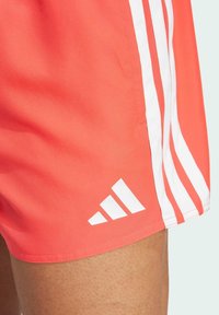 Korálové sportovní šortky s bílými třemi pruhy a logem Adidas. Vyrobeny z lehkého materiálu, s hladkým povrchem.