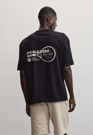 Mand vendt væk, iført en sort Pegador t-shirt med et padelbatsmotiv og beige shorts, stående mod en ensfarvet baggrund.