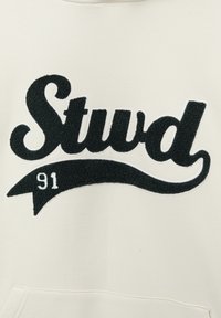 Hoodie crème arborant une grande broderie noire texturée qui épelle "Stwd" avec un design de bannière courbée et "91" en blanc en dessous.