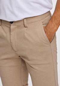 Beżowe lekkie spodnie wykonane z teksturowanej tkaniny, z bocznymi kieszeniami i pojedynczym zapięciem na guzik w pasie.