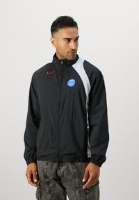 Black nylon jacket with red Nike logo, white side panels, and blue Paris Saint-Germain emblem. Features elastic cuffs and waistband.