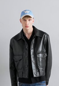 Czarna skórzana kurtka o strukturalnym designie, z dwoma przednimi kieszeniami i zapięciem na guziki. Noszona na czarnej koszuli.