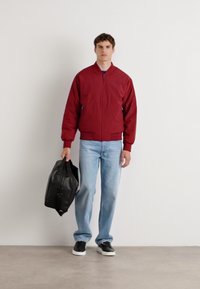 Red bomber jacket with a zip front, blue jeans, and black sneakers. The model holds a black leather bag. Neutral background.