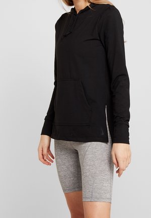 Femme portant un sweat à capuche noir à manches longues avec poche avant et un short gris ajusté au niveau des genoux sur un fond uni.