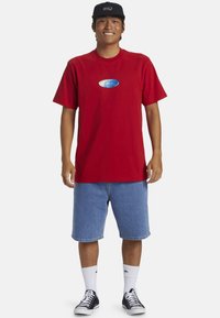 Camiseta de algodón roja con un gráfico azul en el pecho, combinada con pantalones cortos de mezclilla azules, calcetines blancos y zapatillas negras, rematada con una gorra negra.