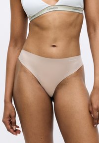 Beige Calvin Klein tanga apakšveļa ar gludu, mīkstu tekstūru. Minimālisma dizains ar zemu jostasvietu un diskreto zīmolu uz jostas.
