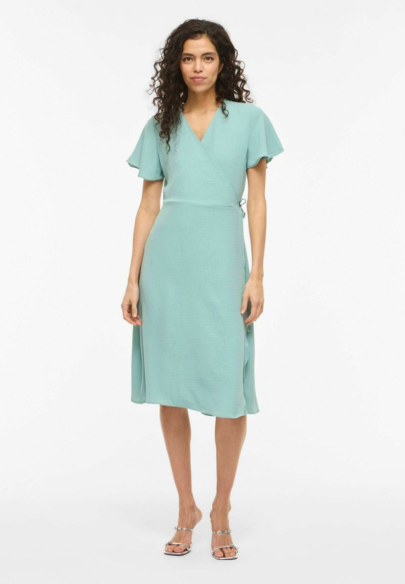 Femme aux cheveux bouclés portant une robe portefeuille bleu clair et des sandales argentées à lanières, debout devant un fond blanc.