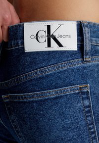 Calvin Klein Jeans, blå denimtyg, figursydd design, synliga sömmar, vit skinnetikett med 'CK'-logotyp och varumärkesnamn på midjebandet.