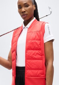 Γυναίκα με λευκή κοντομάνικη μπλούζα Calvin Klein και κόκκινο καπιτονέ γιλέκο κρατώντας ένα γκολφ ρόπαλο στον ώμο μπροστά σε λευκό φόντο.