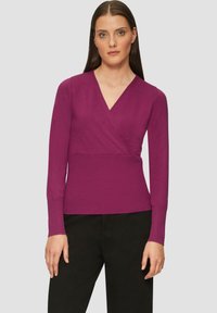 Maglione avvolgente fucsia con scollo a V, maniche lunghe, texture a coste e vestibilità aderente. Indossato con pantaloni neri, stile minimale e tinta unita.