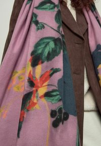 Shawl dengan pola bunga yang menampilkan nuansa merah muda, hijau, kuning, dan biru. Kain yang lembut dan bertekstur disampirkan di atas jaket coklat dengan kerah runcing.