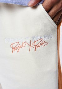 Tejido de crema ligero con bordado en naranja y blanco, que presenta el logo "PROJECT X PARIS" en dos estilos. Detalle del bolsillo visible.