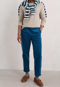 Pantaloni di velluto a coste blu con tasche laterali, abbinati a un maglione di maglia beige e una sciarpa rigata blu e crema annodata attorno al collo. Scarpe marroni.