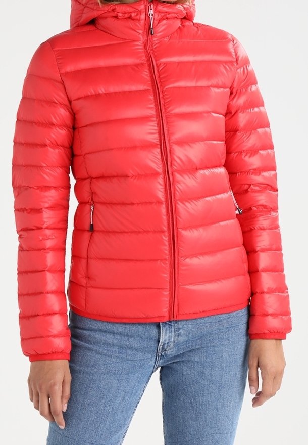 Veste matelassée rouge avec fermeture éclair frontale, poches latérales zippées et capuche attachée portée avec un jean bleu sur une personne, sur fond blanc.