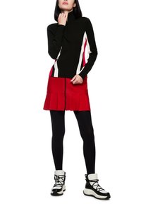 Schwarzes Strickoberteil mit weißen und roten Akzenten, kombiniert mit einem roten plissierten Rock mit einem Reißverschluss vorne. Dazu schwarze Leggings und Stiefel mit weißen Akzenten.