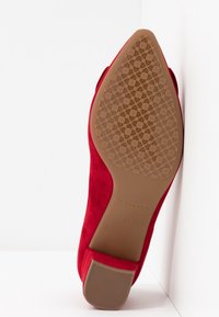 Chaussure à talon aigu en suède rouge avec un bout pointu. La semelle présente un motif floral texturé et une couleur beige. Marque visible sur le talon.