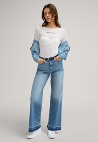 Weißes Mesh-Oberteil mit langen Ärmeln und "JOOP JEANS"-Logo, kombiniert mit hellblauen, hoch geschnittenen, weiten Jeans und weißen Sneakern.