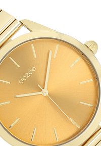 Montre-bracelet dorée avec un cadran métallique lisse présentant de fins marqueurs d'heure et des aiguilles minimalistes. Comprend un bracelet en acier inoxydable doré.