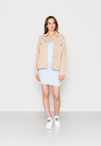 Casaco de ganga bege com bolsos frontais sobre um vestido justo azul claro. A modelo usa ténis brancos com padrões multicoloridos.