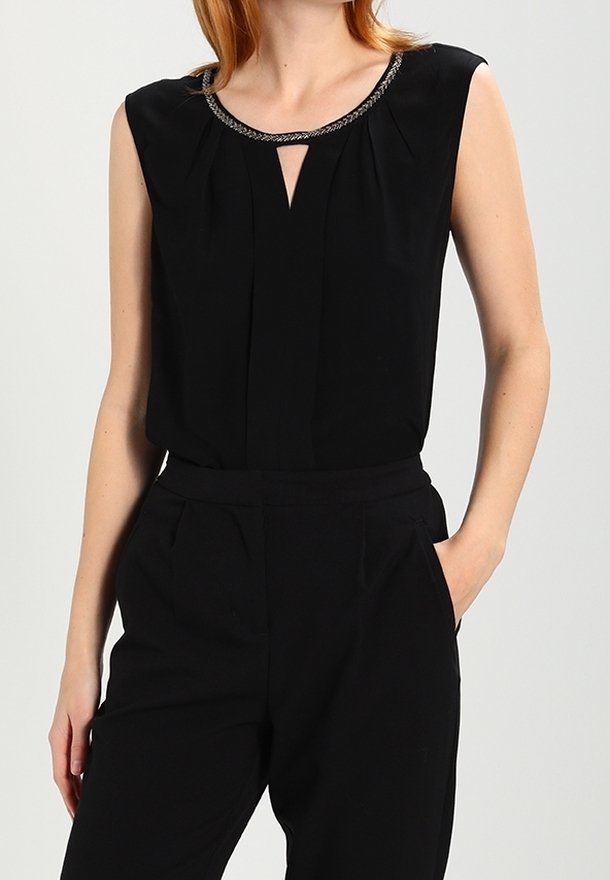 Femme portant un chemisier noir sans manches avec un col décoratif et un pantalon noir taille haute, debout une main dans la poche.