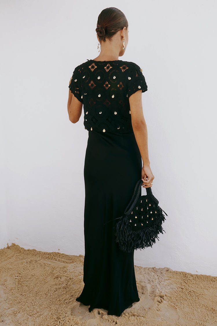 Schwarzes Häkeloberteil mit goldenen Akzenten, kombiniert mit einem langen schwarzen Rock und einer gefransten schwarzen Handtasche. Glatte Textur, figurbetonte Silhouette.