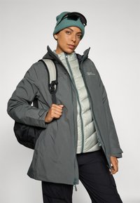 Grau wasserdichte Jacke, die über einer hellgrauen Pufferweste getragen wird, kombiniert mit einer türkisfarbenen Strickmütze und Sonnenbrille, dazu ein schwarzer Rucksack.