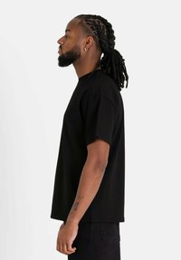 Zwarte katoenen t-shirt met een ontspannen pasvorm, ronde hals en korte mouwen. Het model staat in profiel tegen een neutrale achtergrond.