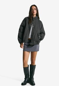 Ženska, oblečena v črno prešito jakno, sivo mini krilo, sivo pulover in črne škornje do kolen, stoji ob belem ozadju.