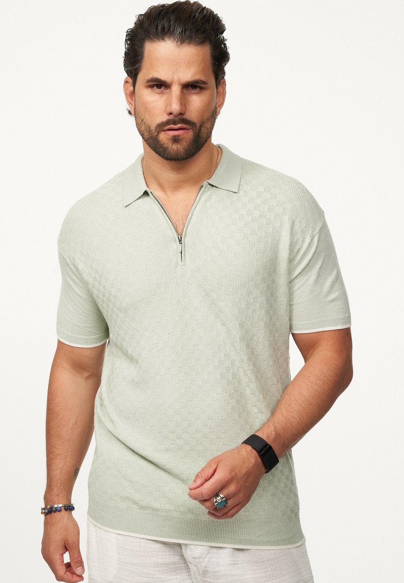 Helles grünes Poloshirt mit kurzem Ärmel und Reißverschlusskragen, strukturiertem Muster, gerippten Bündchen und figurbetonter Passform. Zubehör umfasst Armbänder.