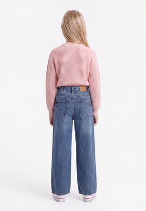 Enfant aux longs cheveux blonds portant un pull rose et un jean bleu à jambes larges, debout de dos sur un fond blanc.