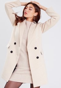 Abrigo beige de doble botonadura con botones negros, bolsillos laterales y mangas amplias, superpuesto a un vestido de suéter beige claro. Textura suave.