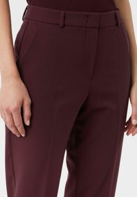 Zbliżenie na osobę w ciemnofioletowych spodniaczkach na miarę z bocznymi kieszeniami i szlufkami na pasek, ręce swobodnie po bokach.