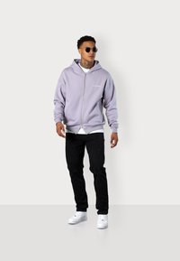 Purple zip-up hoodie with a drawstring hood, paired with black distressed jeans and white sneakers. Short sleeves and minimal branding.
