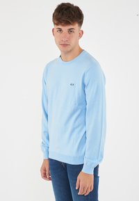 Maglione azzurro chiaro realizzato in tessuto morbido, con maniche lunghe, scollatura a girocollo e un piccolo logo nero sul lato sinistro del petto.