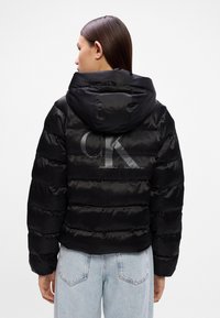 Czarna kurtka puchowa z kapturem, z poziomymi pikowanymi sekcjami. Logo "CK" widoczne w kontrastowym odcieniu na plecach.