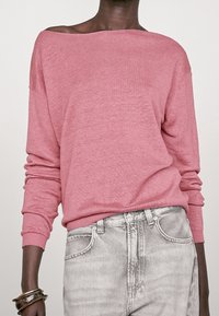 Różowy sweter z odkrytymi ramionami w miękkiej, dzianinowej fakturze, zestawiony z jasnoszarymi jeansami. Sweter ma ściągacze na mankietach i dole.