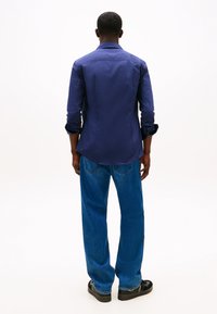 Camisa azul-marinho com botões, textura lisa, mangas compridas enroladas nos punhos, combinada com jeans azuis folgados e sapatos pretos.