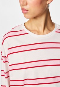 Woman wearing white shirt with horizontal red and pink stripes, silver hoop earring, looking to the side against a plain background.