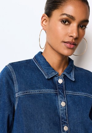 Frau mit glattem, zurückgekämmtem Haar trägt große goldene Creolen und eine blaue Jeansjacke mit sichtbaren weißen Nähten und silbernen Knöpfen.