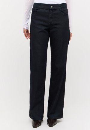Person wearing high-waist black wide-leg jeans paired with dark pointed-toe shoes and a white long-sleeve top, standing against a plain background.