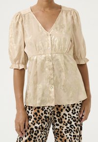 Top oro pallido e lucido con scollo a V e maniche a sbuffo, dotato di vita arricciata e piccoli bottoni. Abbinato a pantaloni con stampa leopardata.