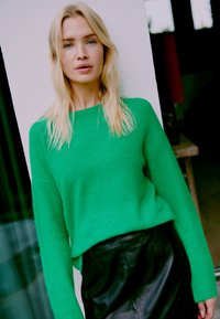 Žalias megztas džemperis su laisvu siluetu. Audinys yra minkštas ir pūkuotas, derinamas su juoda odine sijonu, sukuriant ryškų spalvų kontrastą.