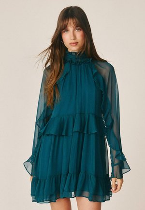 Joven con el pelo largo y castaño lleva un vestido largo de manga larga en color azul verdoso, transparente, con detalles de volantes y cuello alto, sobre un fondo liso.